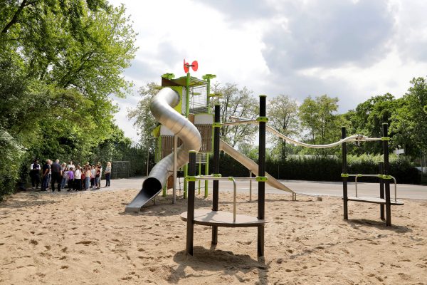 Weitwinkelaufnahme eines Proludic-Spielplatzes mit Kanopé-Turm, Edelstahlrutsche, Seilbahn und Kletterelementen, Gruppe von Personen im Hintergrund