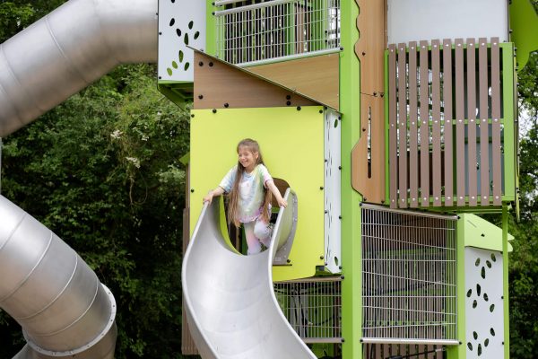 Mädchen rutscht lachend aus dem grünen Proludic-Kanopé-Turm auf einer Edelstahlrutsche