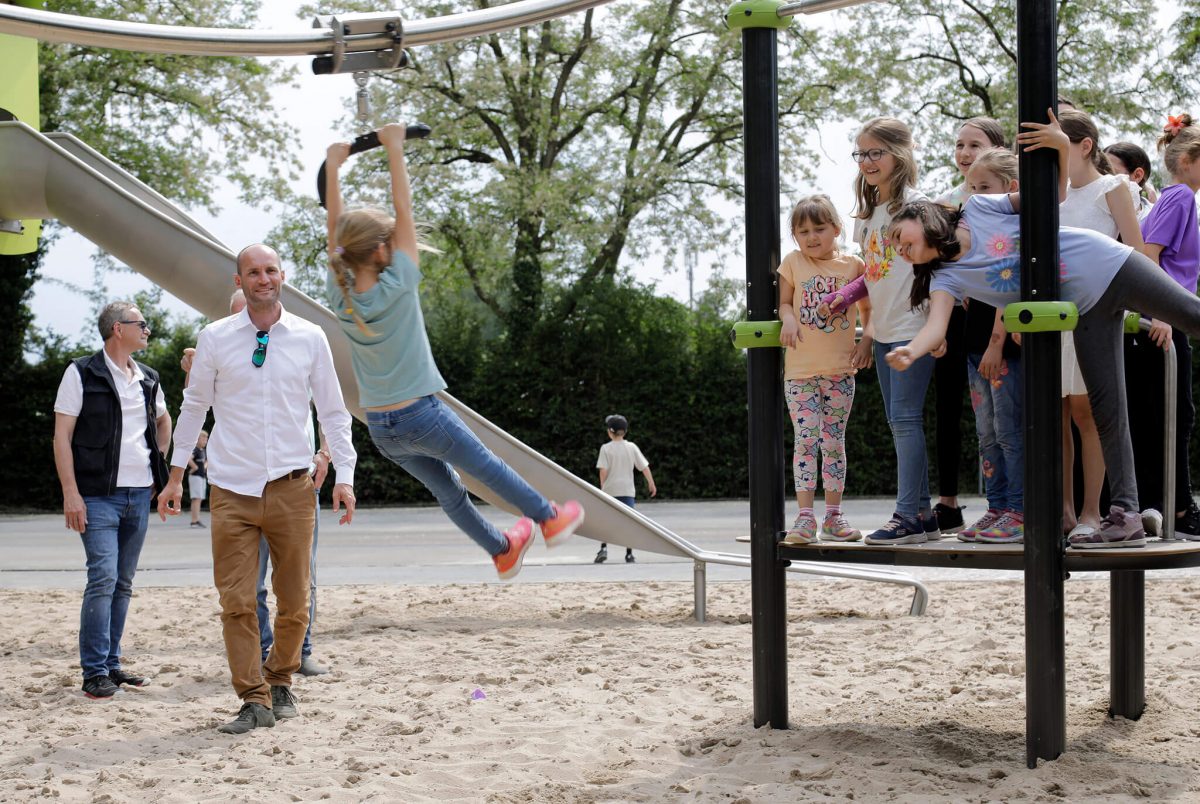 Kinder auf einem Proludic-Spielplatz beim Spielen an einer Seilbahn mit wartenden Kindern und begleitenden Erwachsenen im Hintergrund