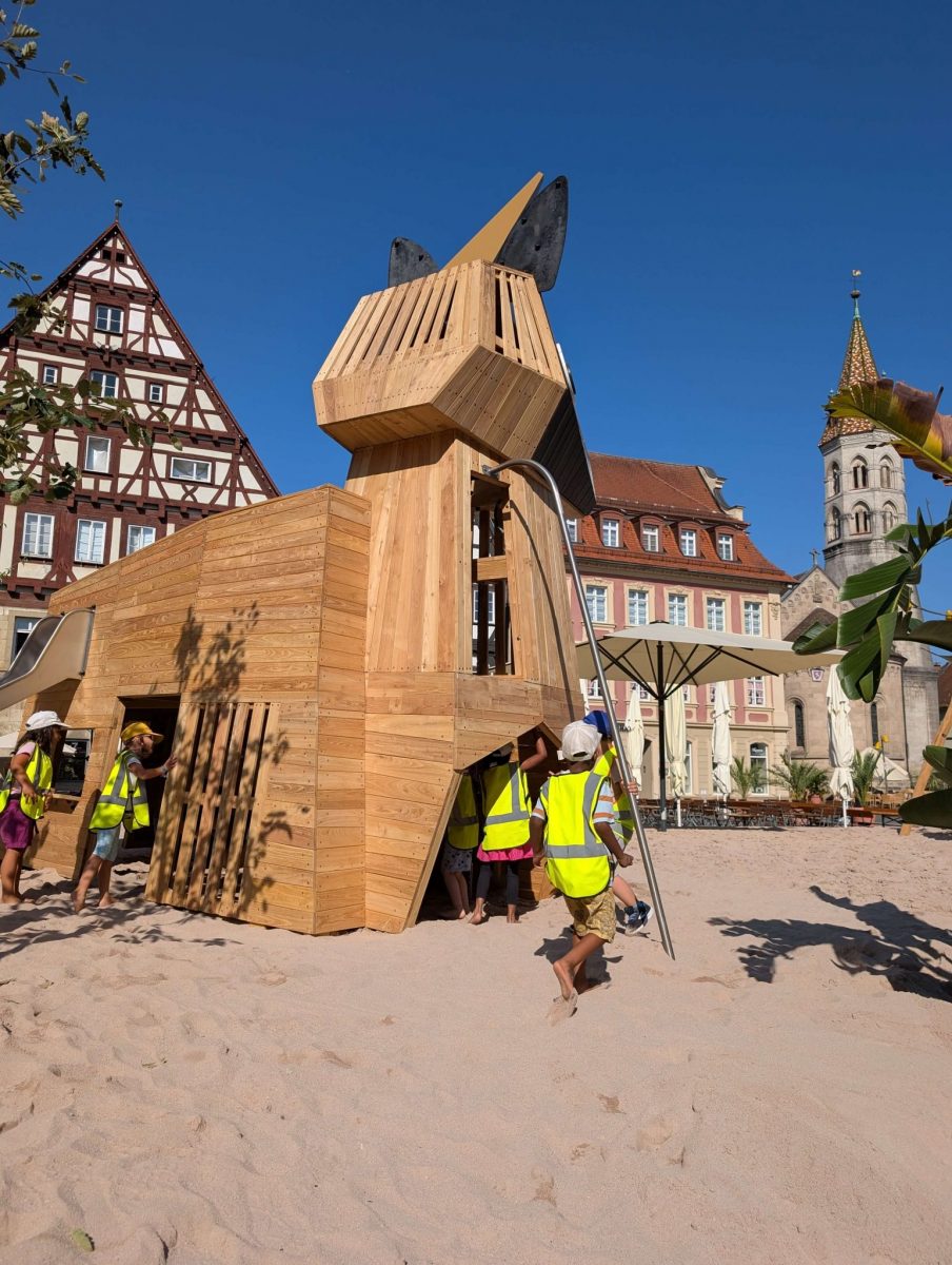 Kinder in Warnwesten bespielen ein großes hölzernes Einhorn-Spielgerät auf Sandfläche. Im Hintergrund sind die historischen Gebäude des Marktplatzes zu sehen. Es ist strahlend blauer Himmel und ein sommerlicher Tag.