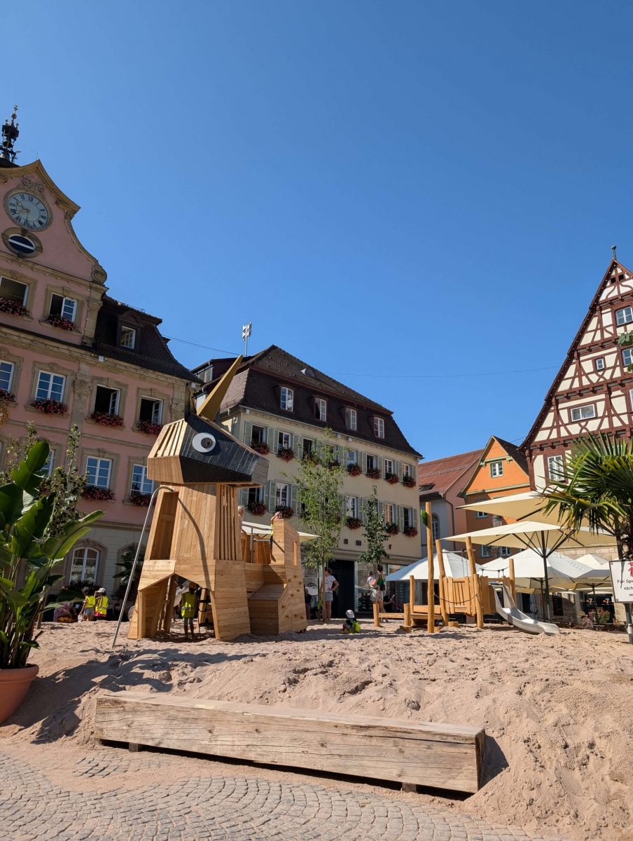 Ein großes, tierförmiges Holzspielgerät mit einem langen, spitzen Horn steht auf einem Sandplatz vor historischen Fachwerkhäusern und einem barocken Gebäude. Im Vordergrund stehen tropische Pflanzen, was den sommerlichen Flair unterstützt. Der Himmel ist wolkenlos und blau.