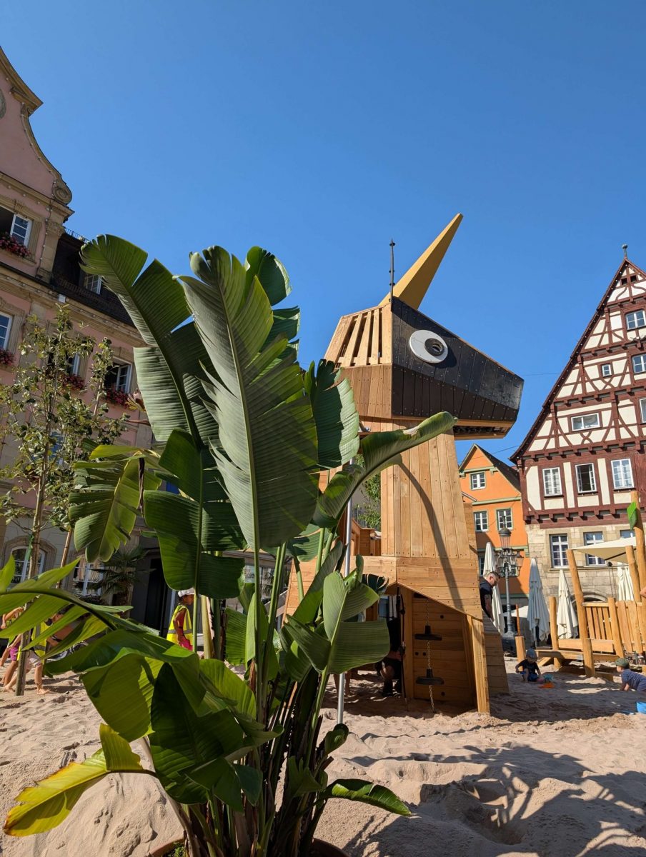 Ein großes, tierförmiges Holzspielgerät mit einem langen, spitzen Horn steht auf einem Sandplatz vor historischen Fachwerkhäusern und einem barocken Gebäude. Im Vordergrund stehen tropische Pflanzen, was den sommerlichen Flair unterstützt. Der Himmel ist wolkenlos und blau – es scheint ein sonniger Tag zu sein beim Einhorn-Spielplatz Schwäbisch Gmünd.