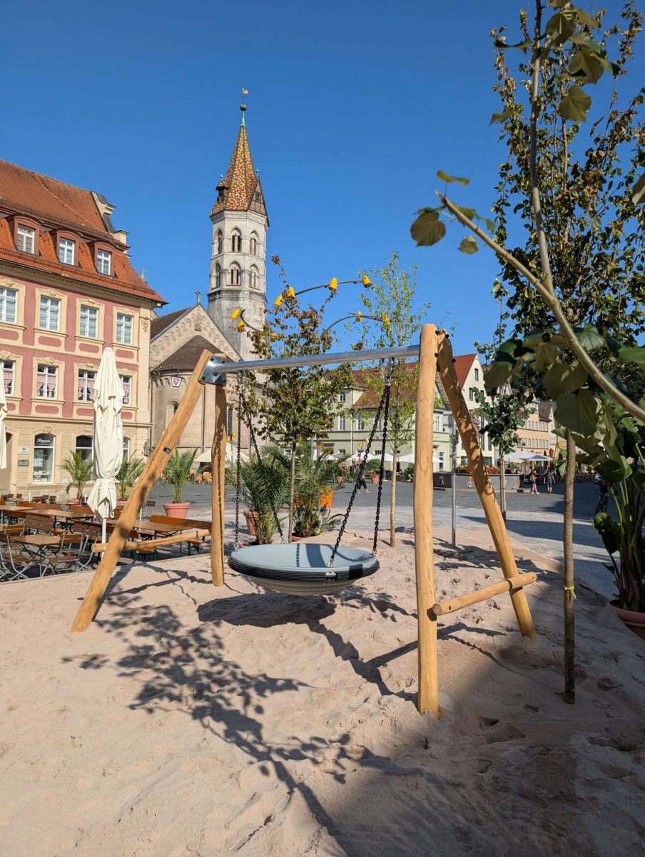 Eine Nestschaukel mit hölzernem Schaukelgestell steht auf dem Sandspielbereich. Im Hintergrund ist die Kirche auf dem Marktplatz von Schwäbisch Gmünd zu sehen. Sonnenschirme und Gastronomiemöbel unterstreichen das gastronomische Angebot rund um den Einhorn-Spielplatz Schwäbisch Gmünd.