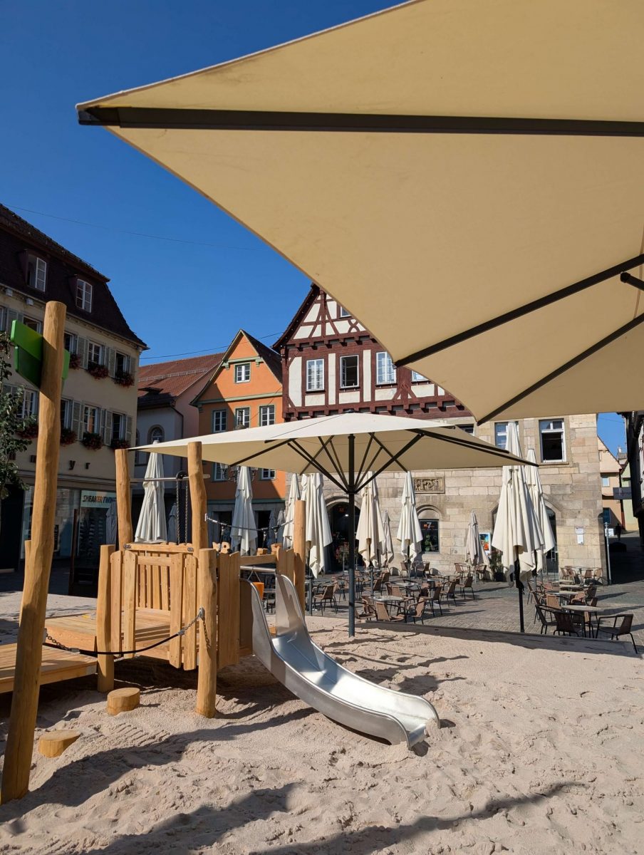 Durch Sonnenschirme wird der Sandspielplatz beschattet. Das Spielgerät mit Rutsche für Kleinkinder steht im Vordergrund. Dahinter sind Sitzmöbel der örtlichen Cafés zu sehen.