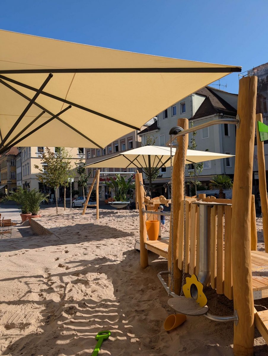 Ein Sandspielgerät für Kleinkinder steht auf einer großen Sandfläche auf dem Marktplatz. Sonnenschirme beschatten die Spielfläche. Das Naturholz der Geräte zeigt die Natürlichkeit des Einhorn-Spielplatz Schwäbisch Gmünd.