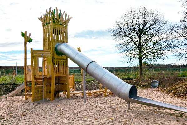 Holz-Kletterturm mit langer Edelstahlrutsche auf einer Sandfläche, umgeben von Natur.