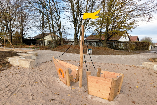 Kleines hölzernes Spielschiff mit gelber Fahne auf einer Sandfläche im Spielplatzbereich.