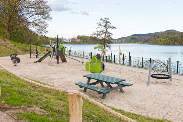 Spielplatz am See mit Rutsche, Kletterelementen, Nestschaukel und Sitzbank auf einer Sandfläche, im Hintergrund der Blick über das Wasser und die umliegenden Hügel.