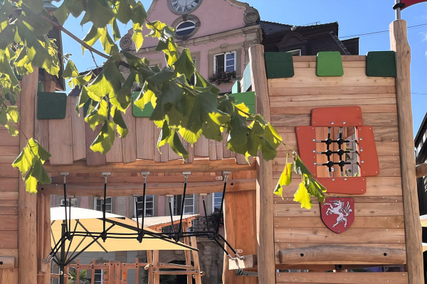 Kinder spielen auf dem Stauferburg Spielplatz in Schwäbisch Gmünd-Rechberg. Das große Holz-Spielschloss mit Türmen, Fenstern und Kletterelementen steht im Sand unter Bäumen, während Erwachsene und weitere Kinder im Hintergrund die Sonne genießen.