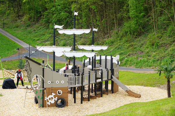 Großes Spielschiff aus Holz und Metall mit Rutsche, Kletternetzen und Spielpodesten, gelegen am Seeufer in naturnaher Umgebung.