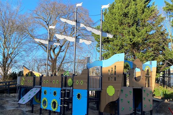 Großes Spielschiff mit bunten Kletter- und Spielelementen, blau-braunen Paneelen, gelben Symbolen, hohen Masten mit weißen Segeln und verschiedenen Aufstiegen.