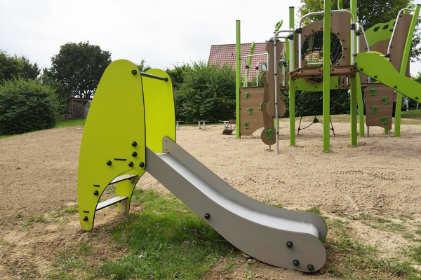 Kleine Rutsche mit grünem Aufstiegselement auf einem Spielplatz, im Hintergrund eine größere Kletter- und Spielanlage mit Netzen und Kletterwänden.