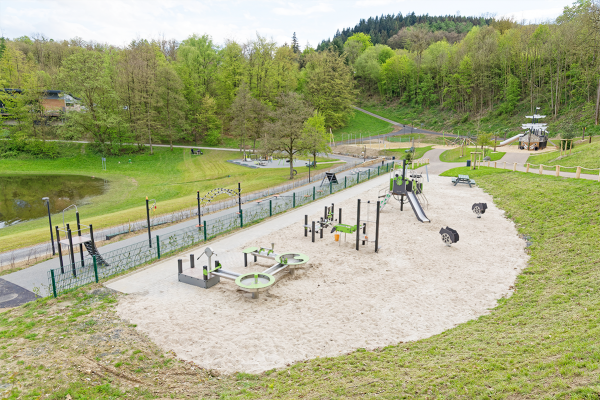 Moderner Spielplatz mit Sandfläche, Klettergeräten, Rutschen und Wasserspielbereich in naturnaher Umgebung mit Wiesen und Wald.