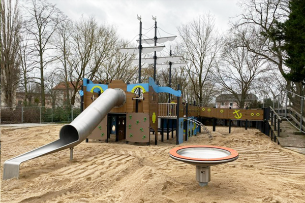 Großes inklusives Piratenschiff-Spielgerät auf einem Spielplatz mit langer Edelstahlrutsche, Kletterwänden, Brücken, hohen Masten mit Segeln und einer Drehscheibe im Vordergrund.