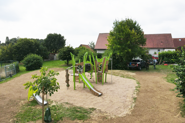 Spielplatz mit grüner Kletter- und Rutschanlage auf einer Sandfläche, inklusive Rutsche, Netzbrücke, Kletterelementen und kleinerer Rutsche im Hintergrund, im Umfeld Bäume und Wohnhäuser.