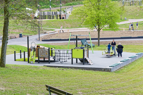 Inklusiver Spiel- und Bewegungsbereich mit barrierefreien Spielgeräten, Schaukeln und Fitnessstationen, genutzt von Kindern und Erwachsenen in naturnaher Umgebung.