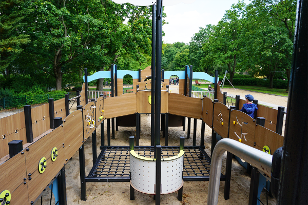 Kinder-Spielplatzgerät in Form eines großen Holzschiffes mit Kletter- und Spielmodulen, Netzen, Spielwänden und Sanduntergrund. Ein Kind spielt an der Seite.