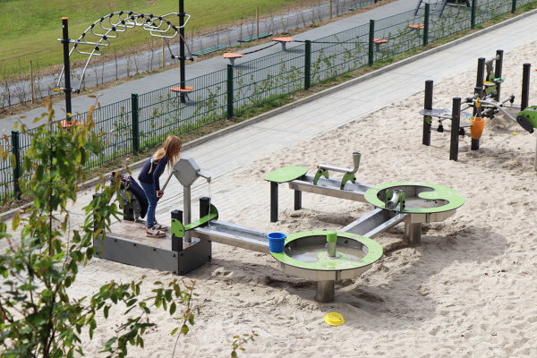 Mädchen spielt an einer Wasserspielanlage mit Pumpstation und Rinnen auf einem Sandspielplatz.
