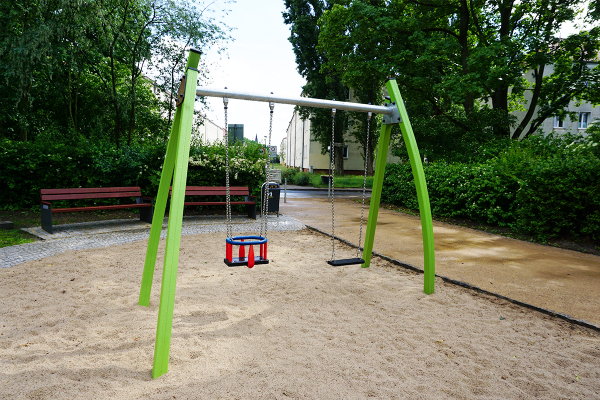 Doppelschaukel auf einem Spielplatz mit einem Kleinkindsitz und einem klassischen Sitz, eingebettet in Sandfläche mit umliegenden Bänken und Grünflächen.