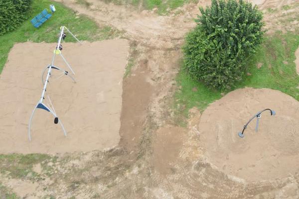 Spielplatz aus der Vogelperspektive mit zwei separaten Bereichen: links eine große Schaukelanlage mit mehreren Sitzen, rechts ein Rundschwinggerät, beide eingebettet in Sandflächen.