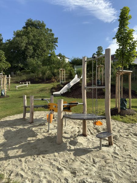 Spielbereich mit Sandfläche, Sandspielanlage und Rutsche in naturnaher Umgebung mit Bäumen und Wiesen.