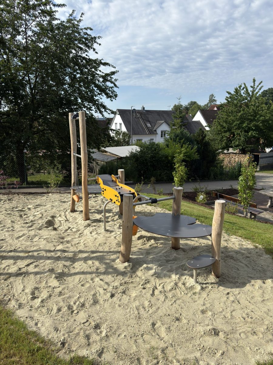 Sandspielbereich mit Sandspielgerät aus Holz und Edelstahl, umgeben von einer naturnahen Umgebung mit Bäumen und Häusern im Hintergrund.