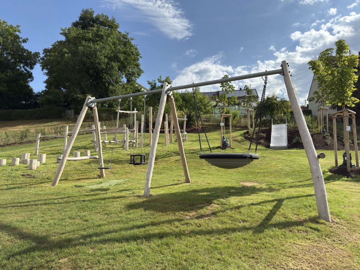Spielplatz im Grünen mit Holzschaukel, Balancier- und Kletterelementen aus Robinienholz sowie Rutsche, eingebettet in eine naturnahe Umgebung.