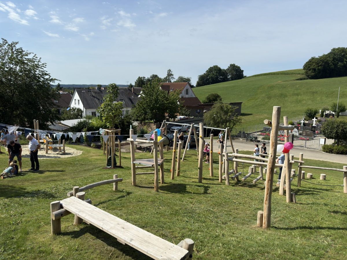 Familien und Kinder spielen auf einem naturnahen Spielplatz mit Kletterelementen aus Robinienholz, Balancierbalken und Sandspielbereich, umgeben von Grünflächen und Dorfkulisse.