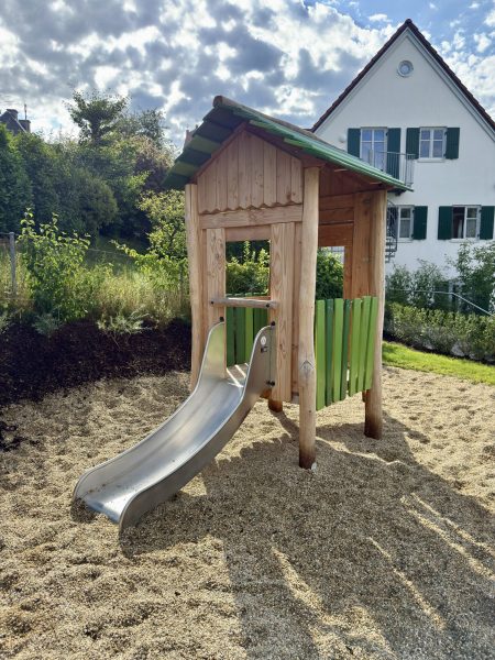 Holz-Spielhaus mit Rutsche auf einem Spielplatz, eingebettet in eine naturnahe Umgebung mit Kiesboden.