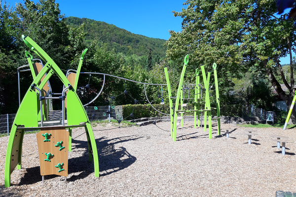 Modernes Kletterspielgerät mit Kletterwand, Seilnetz und Balancierstangen auf einem mit Hackschnitzeln bedeckten Spielplatz, umgeben von Bäumen und Bergen.