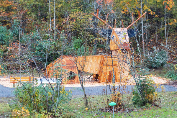 Holzspielskulptur in Form eines Hirsches auf einem Naturspielplatz, umgeben von herbstlichem Wald und Wiese.