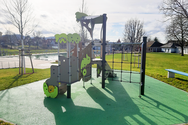 Spielanlage „Abenteuerland“ mit Rutsche, Kletternetz und Spieltafeln auf grünem Fallschutzboden, im Hintergrund Wiese, Bäume und ein Freibadgelände.