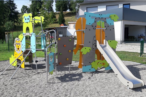 Spielgerät mit bunten Tiermotiven, Rutsche, Kletterwand und Podesten auf Kiesfläche, im Hintergrund Wohnbebauung und Grünfläche.