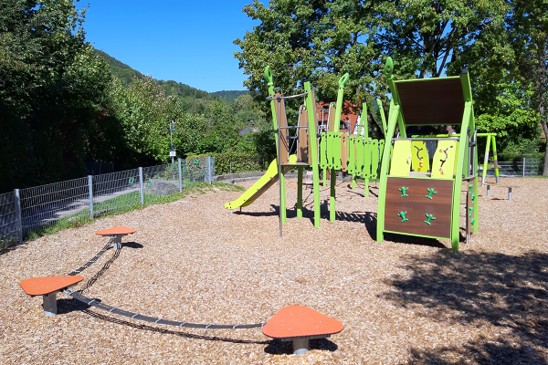 Spielgerät mit Rutsche, Kletterelementen und Hängebrücke auf einem Spielplatz mit Hackschnitzelboden, im Hintergrund Bäume und hügelige Landschaft.