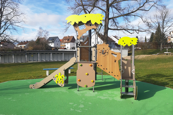 Spielanlage „Abenteuerland“ mit Rutsche, Kletterelementen und bunten Spielelementen auf grünem Fallschutzboden, auf einer Wiese mit Bäumen und Häusern im Hintergrund.