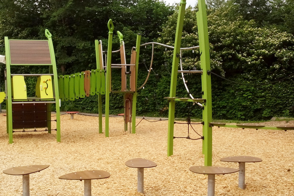 Modulares Kletterspielgerät mit Hängebrücke, Kletternetz und Balancierplattformen auf Hackschnitzelboden, umgeben von dichter grüner Vegetation.