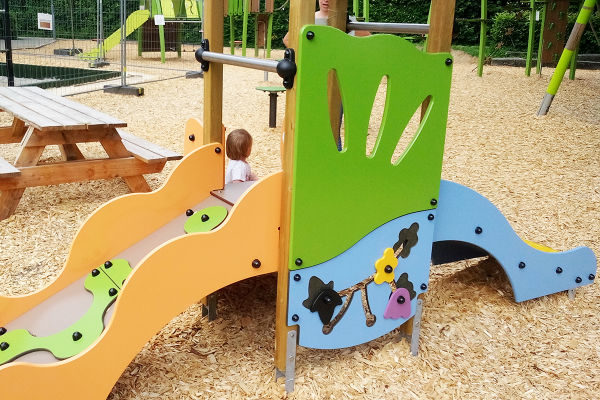 Kleinkindgerechtes Spielgerät mit bunten Farben, Rutsche und taktilen Spielelementen auf Hackschnitzelboden, daneben ein Picknicktisch aus Holz.