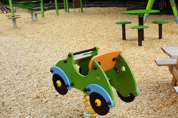 Federspielgerät in Form eines Autos auf Hackschnitzelboden, umgeben von Picknickbank, Balancierstationen und grüner Sitzgruppe.