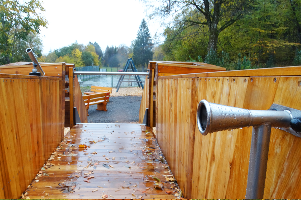 Holzsteg mit Kommunikationsrohren am Hirsch-Spielgerät, Blickrichtung auf den Spielplatz mit Bänken und Bäumen im Hintergrund.