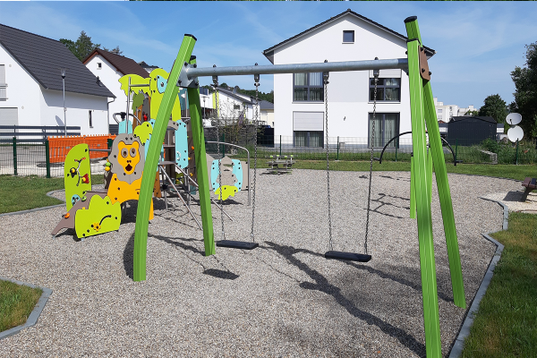 Grüne Doppelschaukel mit zwei schwarzen Sitzflächen auf Kiesfläche, im Hintergrund farbenfrohe Kletterlandschaft und Wohnhäuser.