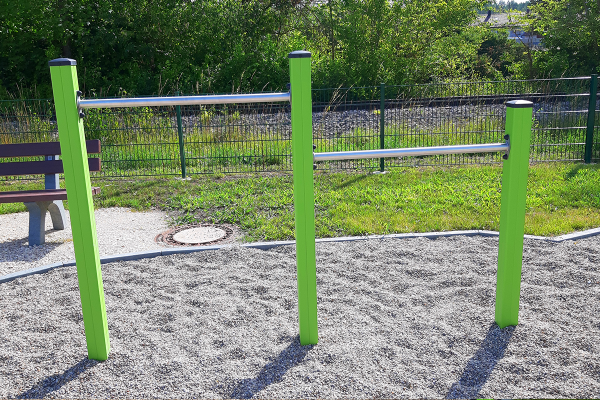 Zwei unterschiedlich hohe Reckstangen mit grünen Pfosten auf Kiesfläche, im Hintergrund eine Bank und Grünfläche.