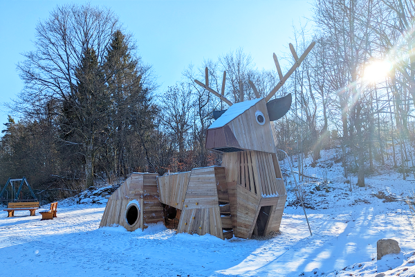 Hirsch-Spielgerät aus Holz auf einem verschneiten Spielplatz mit Sonne und winterlicher Waldkulisse im Hintergrund.