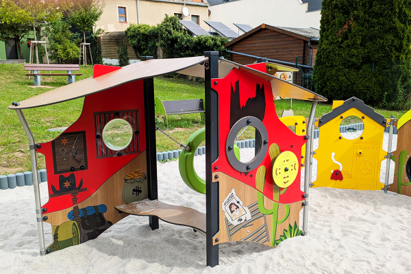 Detailansicht eines thematisch gestalteten Spielmoduls im Western-Stil mit Sheriff-Motiven, Spielgeldsäcken, Steckbrief und runden Sichtfenstern, eingebettet in einen Sandspielbereich.