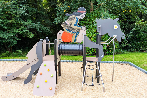 Spielgerät in Form eines Pferdes mit Reiterfigur im Indianer-Stil, ausgestattet mit Kletterelementen, Rutsche und Durchkriechmodul, auf hellem Fallschutzbelag in naturnaher Umgebung.