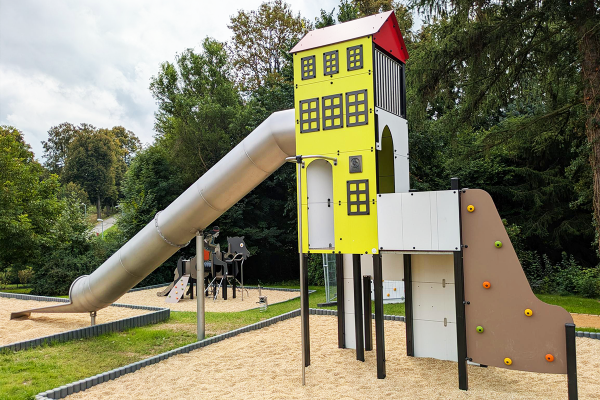 Großer gelber Spielturm mit rotem Dach, langer Edelstahlrutsche und Kletterwand, inspiriert von der Architektur des Karl-May-Hauses, auf sandigem Fallschutzbelag in naturnaher Umgebung.