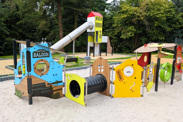 Thematisch gestaltete Spielmodule im Western-Stil mit Motiven wie Saloon, Bank, Tunnelröhre und Sheriff, auf hellem Sandspielboden mit großem Spielturm im Hintergrund.
