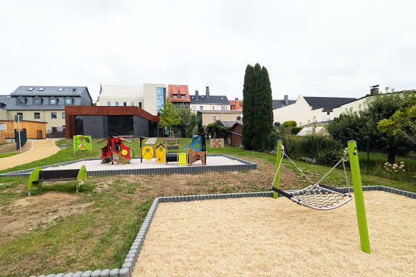 Übersicht des Karl-May-Spielplatzes mit Western-Spielmodulen, moderner Architektur im Hintergrund und einer Netz-Hängematte im Vordergrund auf hellem Fallschutzbelag.