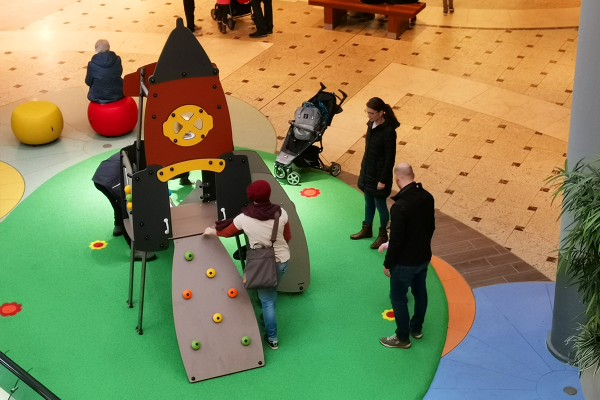 Kinder und Eltern interagieren mit einem Spielgerät in Form einer Rakete auf einer grünen Indoorspielfläche im Einkaufszentrum. Das Gerät verfügt über Kletterelemente und Haltegriffe, der Boden ist mit bunten Blumenmotiven gestaltet.