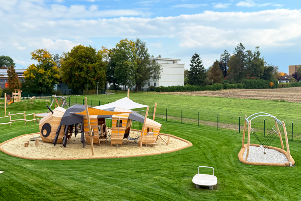 Weitläufiger Spielplatz mit großem Bienen-Spielgerät aus Holz, Sonnensegel, Nestschaukel und Federwippe auf einer grünen Wiese vor ländlicher Kulisse.
