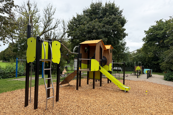 Großes, naturinspiriertes Spielgerät mit Rutsche, Kletterelementen und Verbindungstunnel auf Holzschnitzelboden, im Hintergrund inklusives Spielmodul